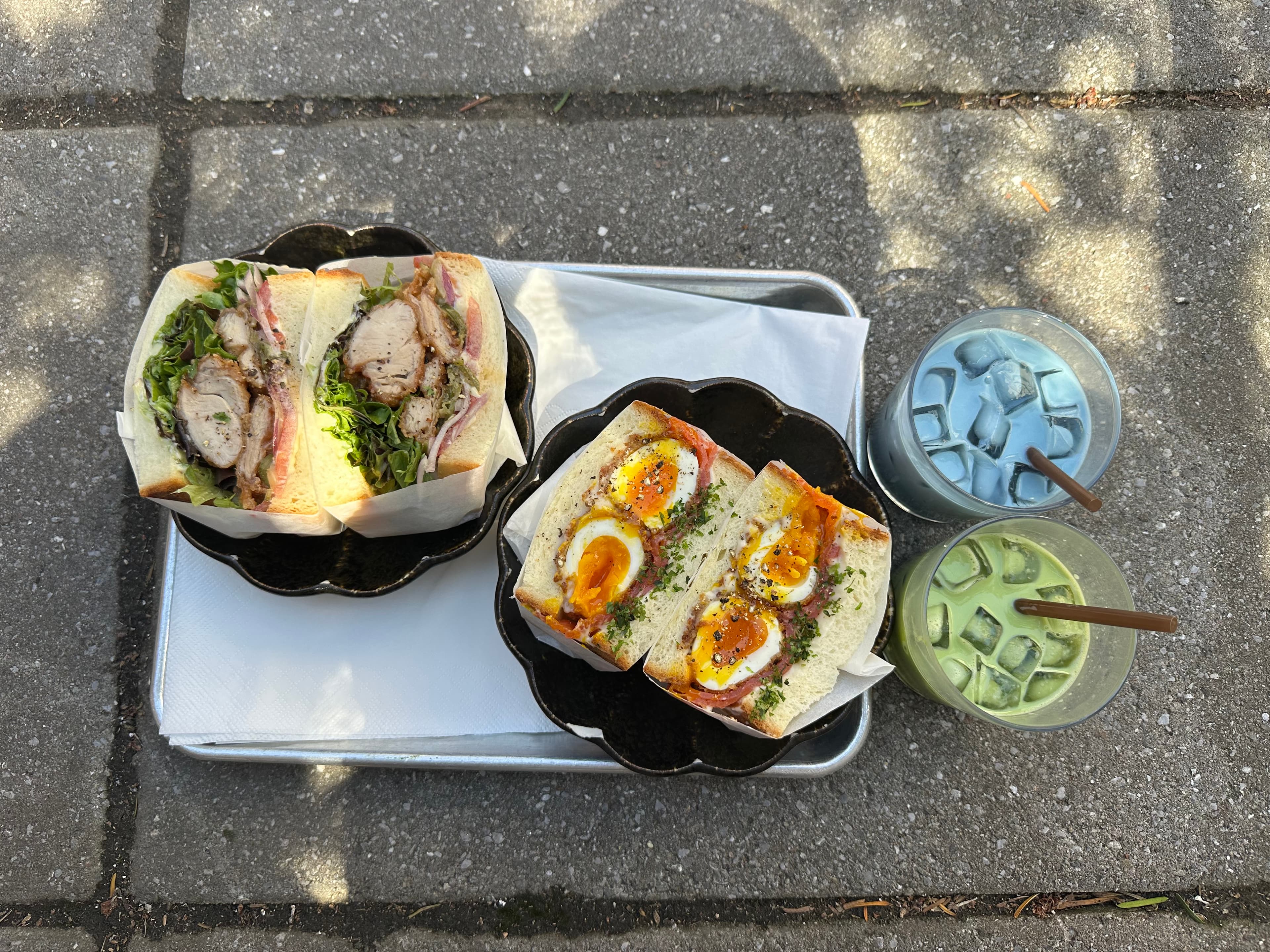 Two sandwiches and iced drinks on a tray, outdoors in the sun.