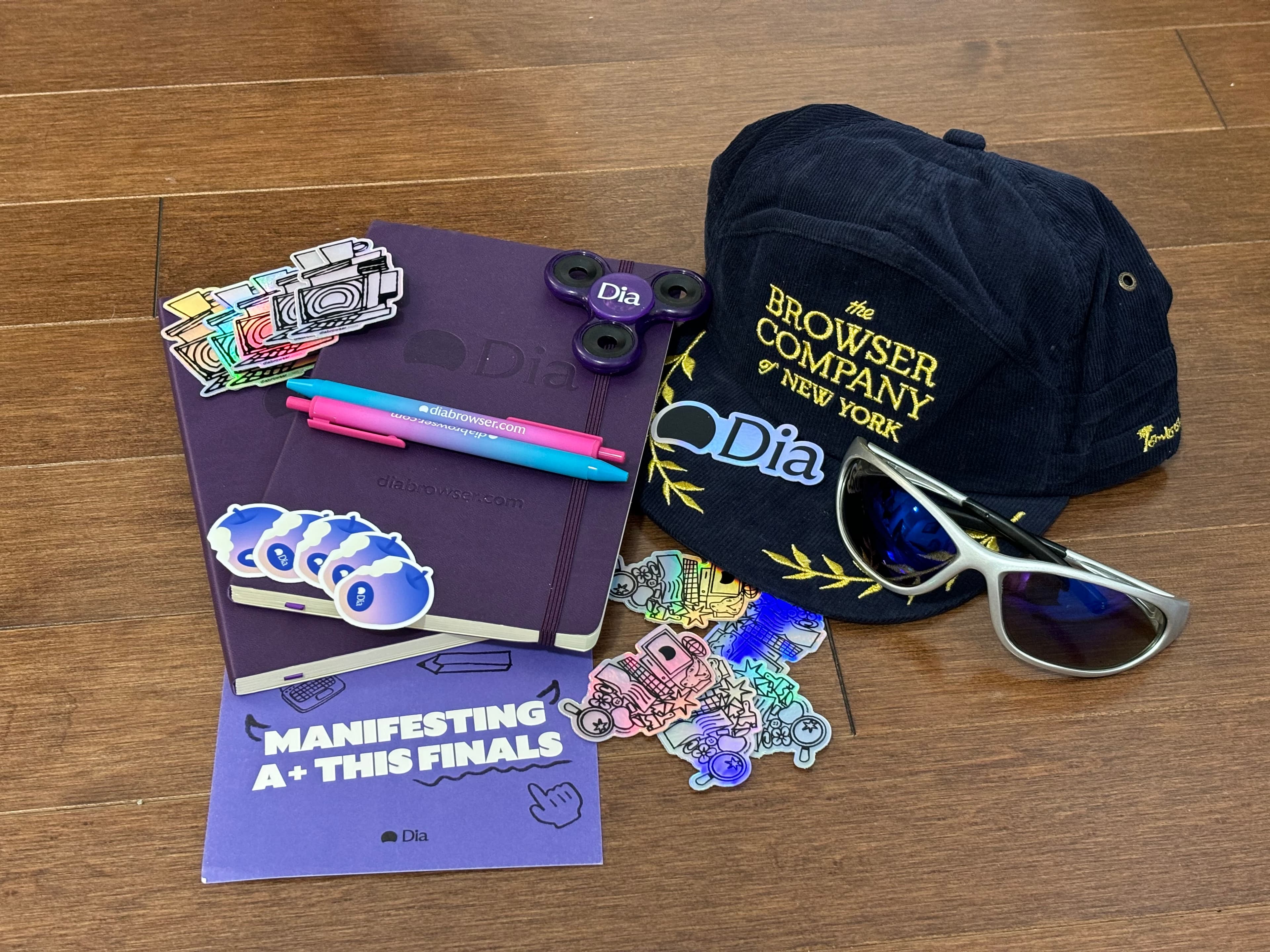 Dia and Browser Company swag laid out on a wood floor: purple notebooks, stickers, pens, a fidget spinner, a corduroy cap and sunglasses.